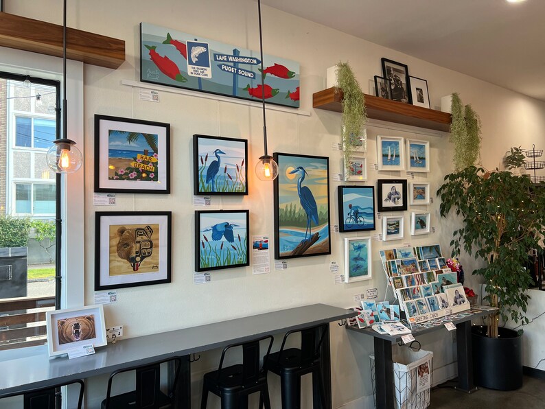K&ouml;nnte beinhalten: Eine Galeriewand mit gerahmten Kunstwerken, darunter Gem&auml;lde von V&ouml;geln, Landschaften und einem B&auml;ren. Die Kunstwerke sind an einer wei&szlig;en Wand mit einem Holzregal dar&uuml;ber angebracht. Auf einem Tisch im Vordergrund befinden sich auch einige Postkarten und Gru&szlig;karten.