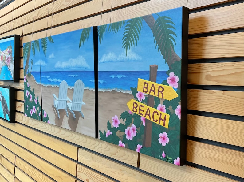 May include: A three-piece beach scene painting set. The paintings feature a blue ocean, white sand, palm trees, and two white chairs. The third painting features a sign that says "Bar" and "Beach" with pink flowers.