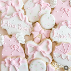 May include: An assortment of decorated sugar cookies in shades of pink and white. The cookies are shaped like bows, dresses, and a baby carriage. Some cookies have the name "Aria Rose" written on them. The cookies are decorated with pink icing and floral patterns.