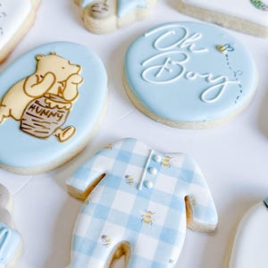 May include: Assortment of baby shower cookies. Includes a blue onesie cookie with a gingham pattern and bee accents, and cookies with Winnie the Pooh and "Oh Boy" designs. The cookies are decorated with blue, yellow, and white icing.