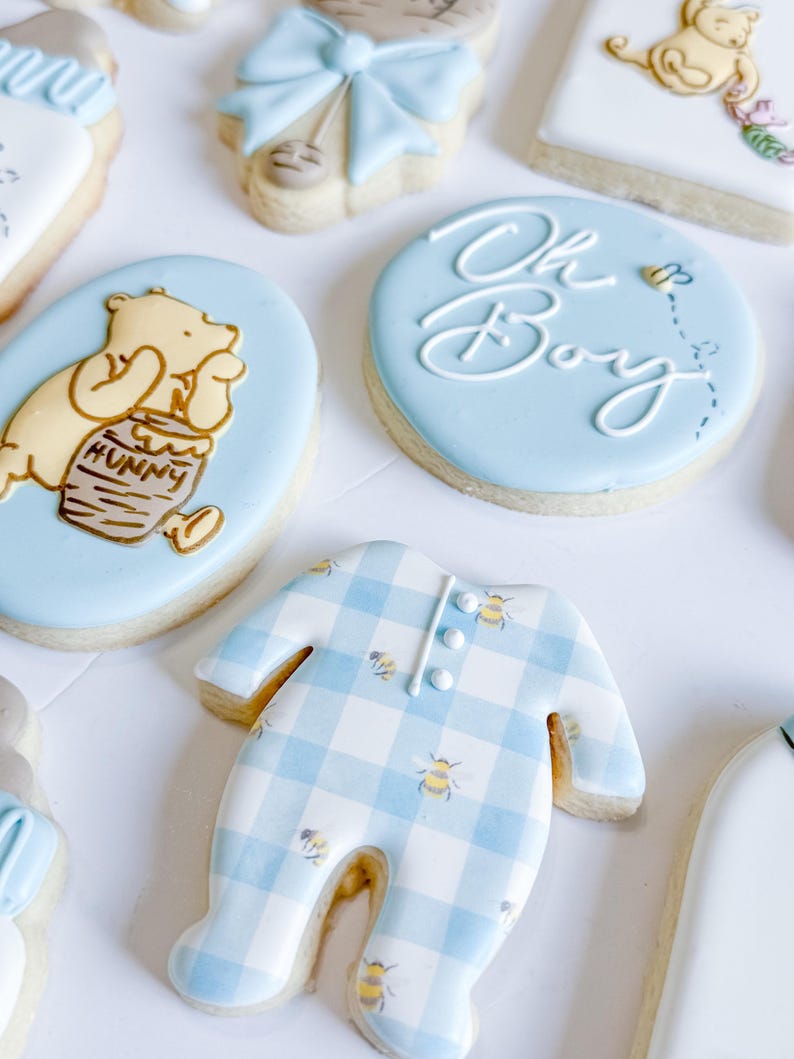 May include: Assortment of baby shower cookies. Includes a blue onesie cookie with a gingham pattern and bee accents, and cookies with Winnie the Pooh and "Oh Boy" designs. The cookies are decorated with blue, yellow, and white icing.