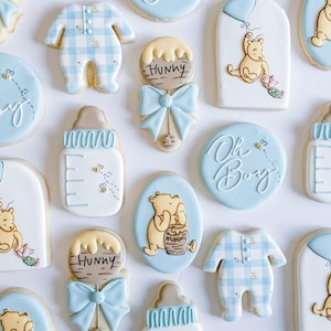 May include: An assortment of baby shower cookies in various shapes and designs. The cookies feature a light blue and white color scheme, with designs including Winnie the Pooh, baby bottles, onesies, and the phrase "Oh Boy".