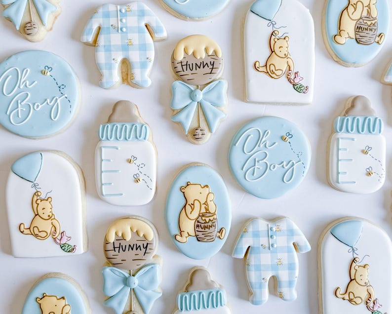 May include: An assortment of baby shower cookies in various shapes and designs. The cookies feature a light blue and white color scheme, with designs including Winnie the Pooh, baby bottles, onesies, and the phrase "Oh Boy".