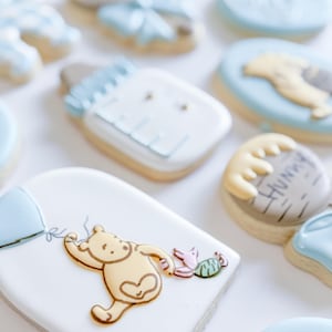 May include: Assortment of decorated sugar cookies with a baby shower theme. Cookies include a baby bottle, Winnie the Pooh with a balloon, and other shapes with blue and yellow icing. The cookies are on a white surface.