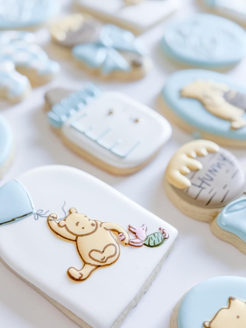 May include: Assortment of decorated sugar cookies with a baby shower theme. Cookies include a baby bottle, Winnie the Pooh with a balloon, and other shapes with blue and yellow icing. The cookies are on a white surface.