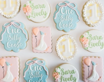 May include: Assortment of decorated sugar cookies with a birthday theme. Cookies include the text "One Silly Goose" and "Sara Lively", along with a goose wearing a party hat and a number one. Colors include pink, blue, white, and yellow.