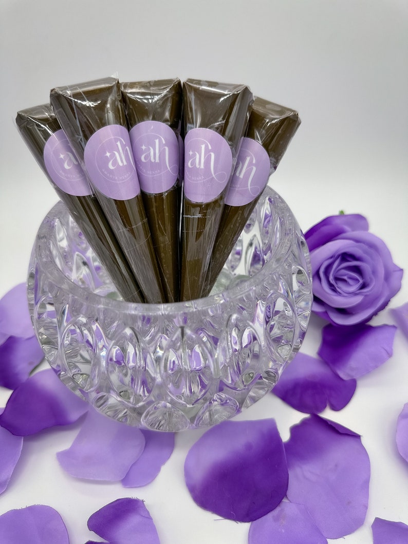 Natural henna cones with purple labels that say 'amarya henna' in white lettering. The cones are in a clear glass bowl with a textured surface. The bowl is surrounded by purple flower petals.