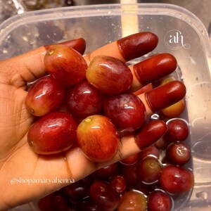 Woman hand with deep red-stained fingertips holding matching red grapes. Made with Amarya Henna Cones