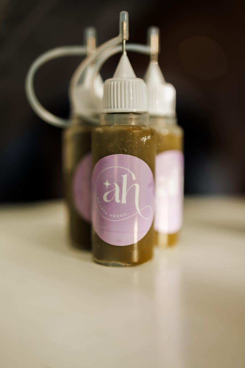 Three small bottles of Amarya Henna freckle bottles with a purple label featuring 'ah' logo on a blurred background