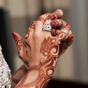 Bride hands adorned with intricate deep red henna designs, complemented by rings, bracelets, and other jewelry, with hands gently clasped together.
