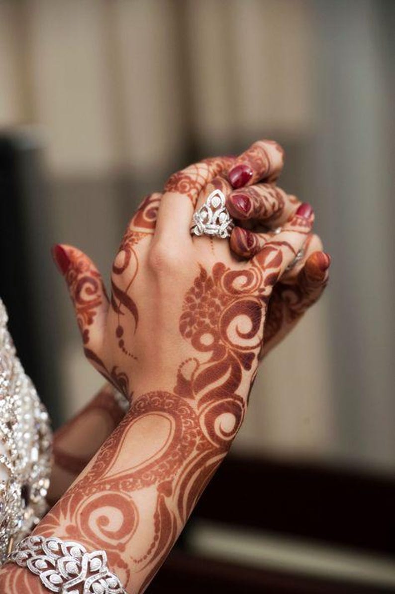 Bride hands adorned with intricate deep red henna designs, complemented by rings, bracelets, and other jewelry, with hands gently clasped together.