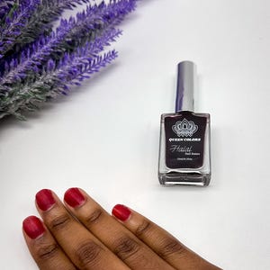 May include: A bottle of Queen Colors Halal Nail Stain in a deep red color. The bottle is clear and has a silver cap. The bottle is sitting on a white surface. A hand with red nail polish is shown in the image.