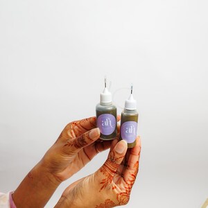Close-up of henna-stained hands holding two Amarya Henna freckles bottle with a lavender-purple label on a white background.