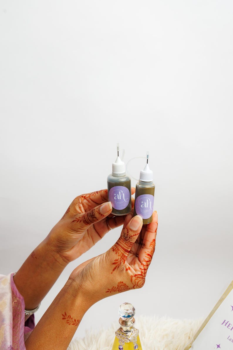 Close-up of henna-stained hands holding two Amarya Henna freckles bottle with a lavender-purple label on a white background.