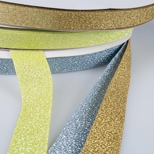 May include: Three rolls of glitter ribbon in gold, lime green, and light blue. Each ribbon features a dense, sparkling texture on the front surface, wound onto circular spools with white cardboard centers, displayed in a stacked arrangement.