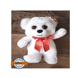May include: White plush teddy bear with large brown eyes and a red satin bow. The bear is standing on a wooden surface.
