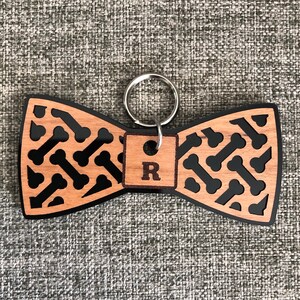 May include: Wooden bow tie keychain with a black bone pattern and a personalized tag with the letter "R" in black.