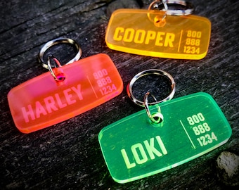 May include: Three brightly coloured plastic dog tags with rounded corners and silver key rings. The tags are orange, yellow, and green. The tags have the names "Harley", "Cooper", and "Loki" engraved on them, along with a phone number.