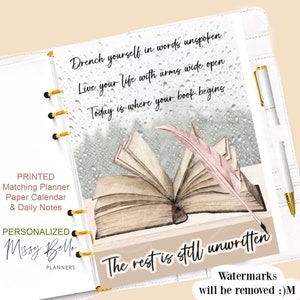 May include: A white planner with a gold coil binding. The cover features an illustration of an open book with a pink feather quill. The text on the cover reads "Drench yourself in words unspoken, Live your life with arms wide open, Today is where your book begins, The rest is still unwritten".