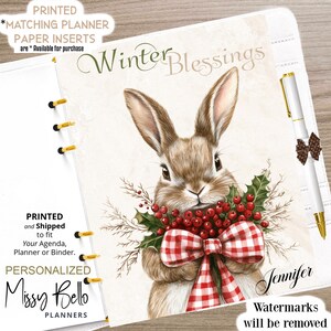May include: A personalized planner with a winter theme. The cover features a brown bunny holding holly and a red and white checkered bow. The text "Winter Blessings" is displayed above the bunny. A pen with a decorative bow is also visible.
