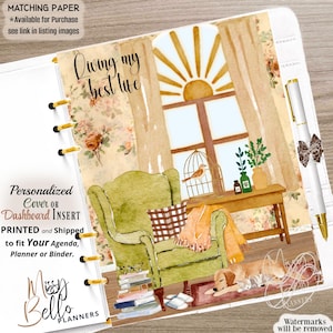 May include: A planner dashboard insert featuring a watercolor illustration of a cozy room with a green armchair, a sunlit window, a dog, and a cat. The text reads Living my best life. The insert includes gold binder rings and a white pen with a bow.