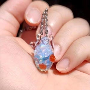 May include: A delicate silver chain necklace with a glass pendant featuring a blue and iridescent design. The pendant has red glass roses attached to it.
