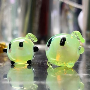 May include: Two small, rounded, light green glass sculptures with black accents and small appendages. The sculptures have a reflective surface and are sitting on a reflective surface. The text "Galaxy M BambooTreats" is visible on the surface.