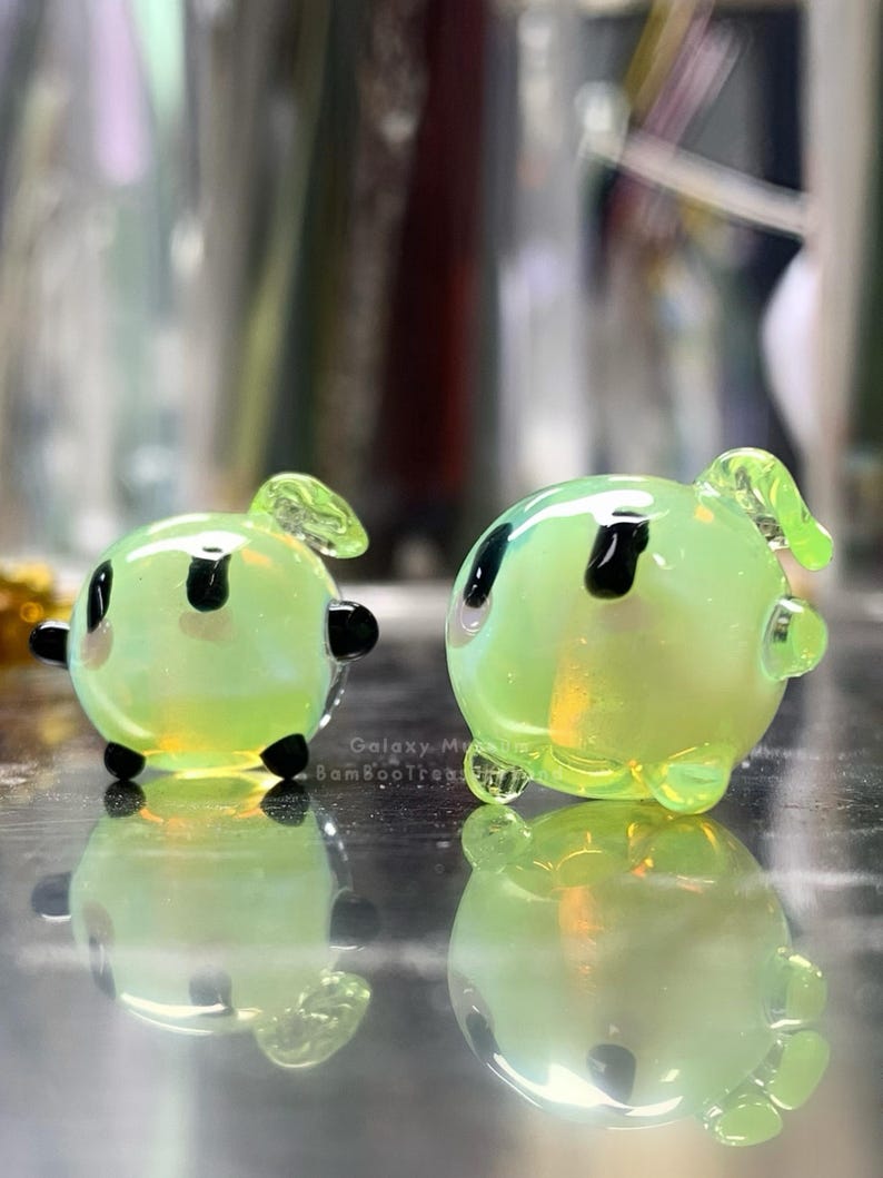 May include: Two small, rounded, light green glass sculptures with black accents and small appendages. The sculptures have a reflective surface and are sitting on a reflective surface. The text "Galaxy M BambooTreats" is visible on the surface.