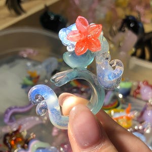 Puede incluir: Anillo de vidrio artesanal en tonos azules y transparentes. El anillo presenta un diseño de caballito de mar estilizado con un acento de flor roja. El vidrio tiene una calidad iridiscente. El anillo es sostenido por una mano. Talla M.