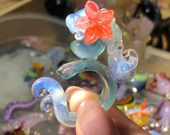 May include: A whimsical, handcrafted glass ring in shades of blue and clear. The ring features a stylized seahorse design with a vibrant red flower accent. The glass has a shimmering, iridescent quality, and the ring is held by a hand.
