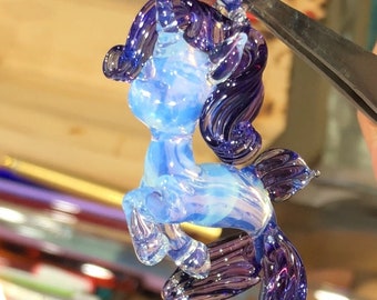 May include: A glass unicorn figurine with a blue and purple iridescent finish. The unicorn has a flowing mane and tail.