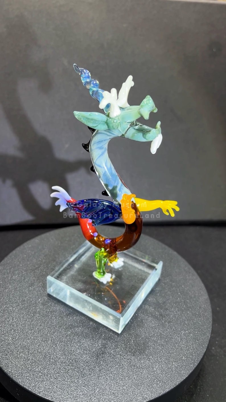 May include: A colorful glass dragon figurine. The dragon features a blue, green, yellow, red, and brown body with white accents. It is mounted on a clear, square base. The text "galaxy BambooTreasureland" is visible on the dragon.