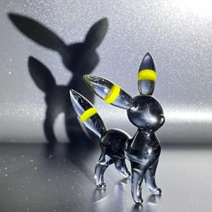 May include: A clear glass figurine of a creature with large ears and a tail, featuring yellow bands on the ears and tail. The figurine casts a shadow on a reflective surface, creating a silhouette of the same form.