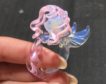 May include: A glass ring with a pink and clear design. The ring features a mermaid tail and a pink swirl design. The ring is made of glass and has a delicate, whimsical look.