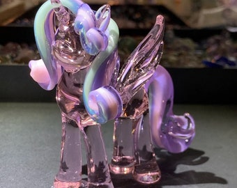 May include: A glass unicorn figurine with iridescent purple, pink, and blue wings and mane. The unicorn is standing on a gray surface.