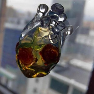 May include: A clear glass heart-shaped ornament with a yellow and green interior and red accents. The ornament has a textured surface and appears to be handmade.