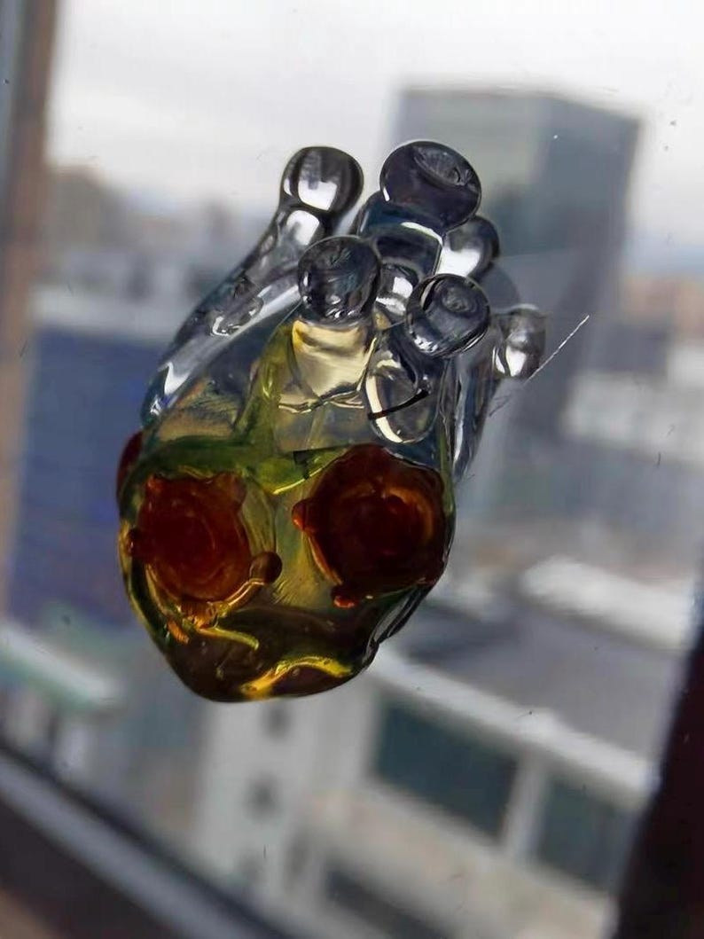 May include: A clear glass heart-shaped ornament with a yellow and green interior and red accents. The ornament has a textured surface and appears to be handmade.