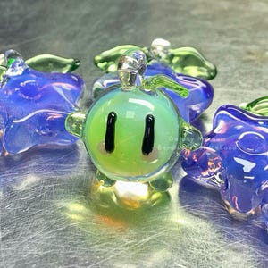 May include: A selection of vibrant glass ornaments. The central piece is a green, spherical figure with black eyes and a smiling mouth. It is surrounded by blue star-shaped and green leaf-shaped glass ornaments. The glass has a shiny finish.