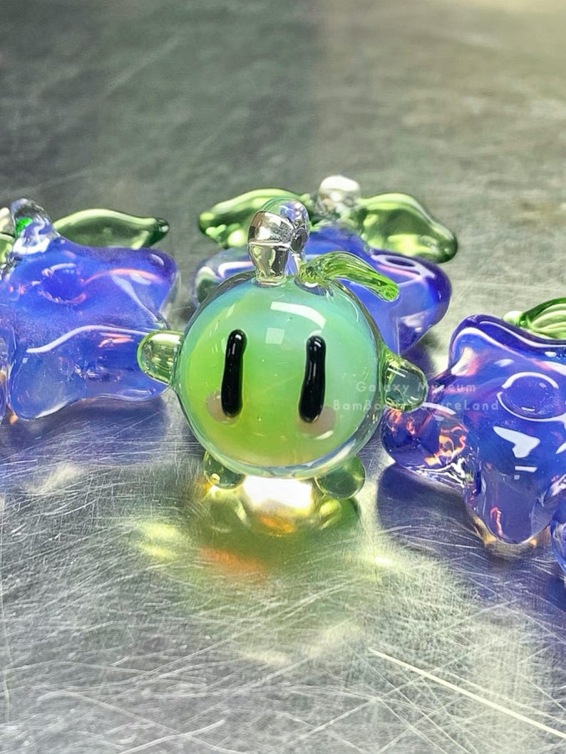 May include: A selection of vibrant glass ornaments. The central piece is a green, spherical figure with black eyes and a smiling mouth. It is surrounded by blue star-shaped and green leaf-shaped glass ornaments. The glass has a shiny finish.