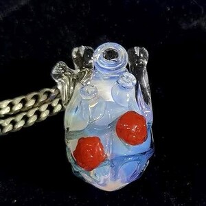 May include: A glass heart pendant with a light blue and white swirl design. The heart has two red glass beads embedded in it. The pendant is attached to a silver chain.