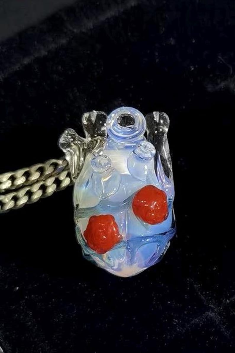 May include: A glass heart pendant with a light blue and white swirl design. The heart has two red glass beads embedded in it. The pendant is attached to a silver chain.