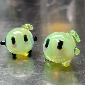 May include: Two small, round, green glass figurines with black eyes, black feet, and small green leaf-like details on top. The figurines have a slight iridescent sheen. The text "Galaxy Museum Bamboo TreasureLand" is visible.