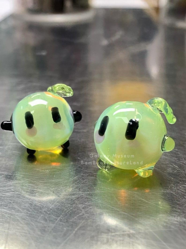 May include: Two small, round, green glass figurines with black eyes, black feet, and small green leaf-like details on top. The figurines have a slight iridescent sheen. The text "Galaxy Museum Bamboo TreasureLand" is visible.
