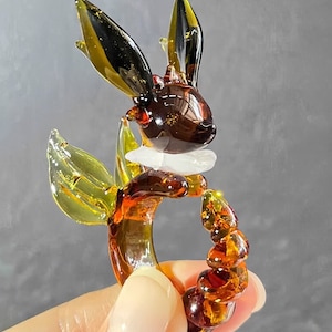 Puede incluir: Un anillo de vidrio con una figurita de conejo marrón y amarillo. El conejo tiene orejas grandes y un collar blanco. El anillo está hecho de cuentas de vidrio.