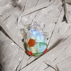 May include: A glass pendant with a clear, iridescent body and red and green accents. The pendant is shaped like a heart and has a delicate, intricate design.