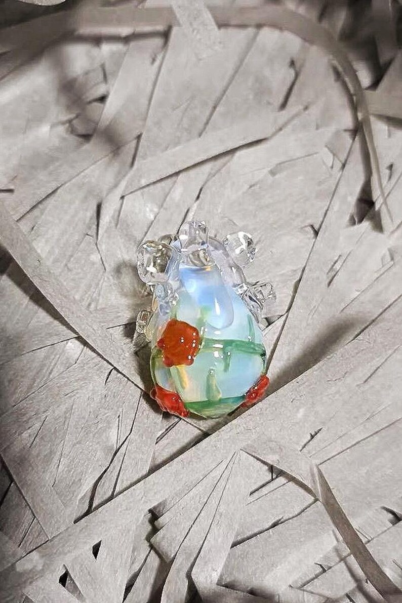 May include: A glass pendant with a clear, iridescent body and red and green accents. The pendant is shaped like a heart and has a delicate, intricate design.