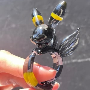 May include: A black glass ring with a yellow stripe design, shaped like a cartoon character with large ears.