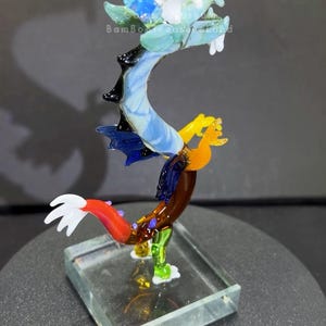 May include: A colorful glass dragon sculpture with a clear base. The dragon features a blue, white, green, yellow, and red design. The sculpture is displayed on a dark surface. The text "Bambo Treasures" is visible.