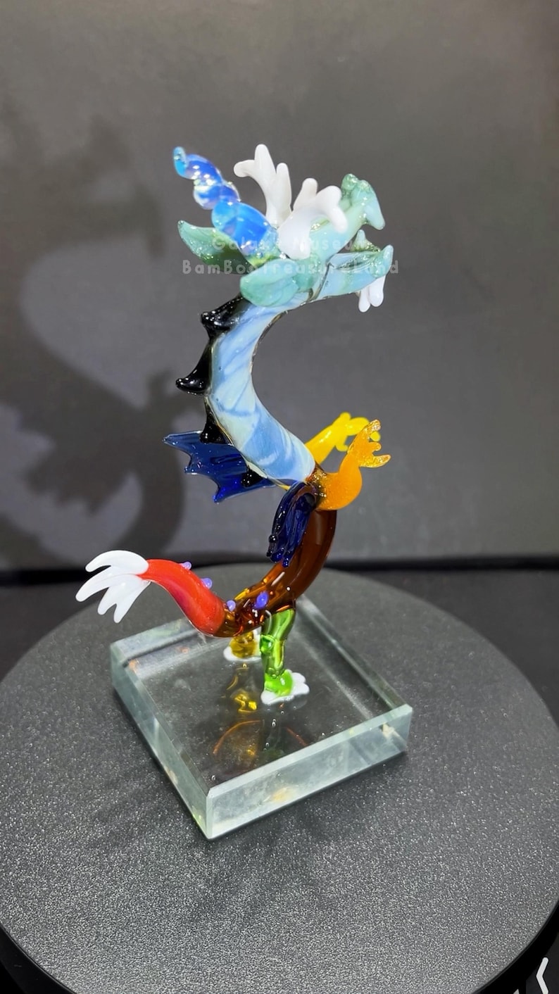 May include: A colorful glass dragon sculpture with a clear base. The dragon features a blue, white, green, yellow, and red design. The sculpture is displayed on a dark surface. The text "Bambo Treasures" is visible.