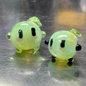 May include: Two small, round, glass figurines in a light green colour. Each has black eyes, pink cheeks, and small black arms and legs. One has a small green leaf on top. The other has a small green leaf on top.